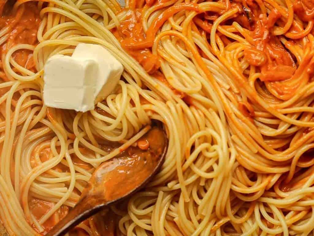 A close-up of cooked spaghetti mixed with a creamy tomato sauce. A wooden spoon stirs the pasta, and two pats of butter rest on top, partially melting into the noodles.